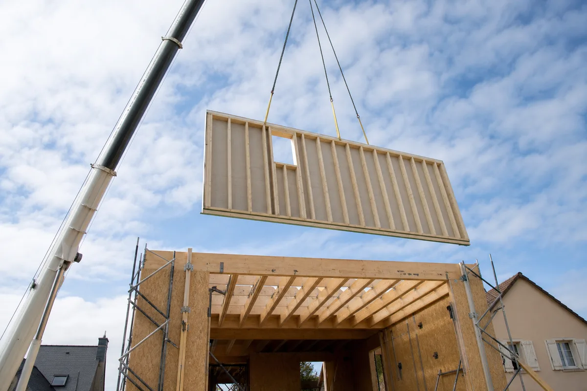 Panneau de mur à ossature bois préfabriqué levé par une grue au-dessus d’une maison sans toiture